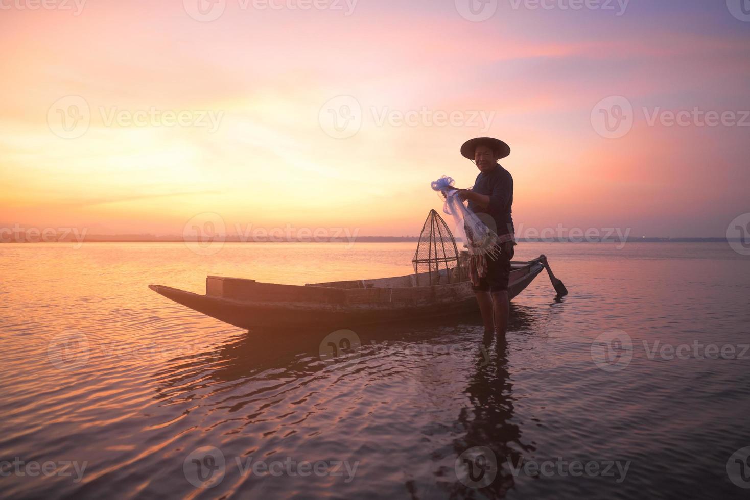 asiatisk fiskare med sin träbåt som håller ett nät redo för att fånga sötvattensfisk i naturfloden tidigt under soluppgången foto