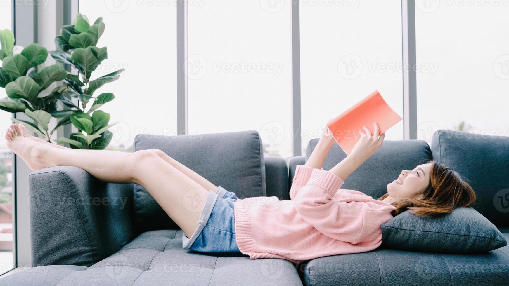 ung asiatisk kvinna läser bok på semester. en närbild av ung glad flicka liggande öppnad bok på soffan hemma. avslappnad livsstil hemma. foto