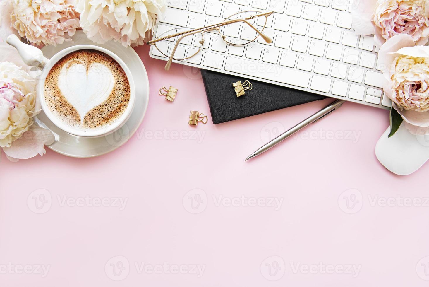 platt låg ovanifrån kvinnors skrivbord med blommor. kvinnlig arbetsyta med laptop, blommor pioner, tillbehör, anteckningsbok, glasögon, kopp kaffe på rosa bakgrund. foto