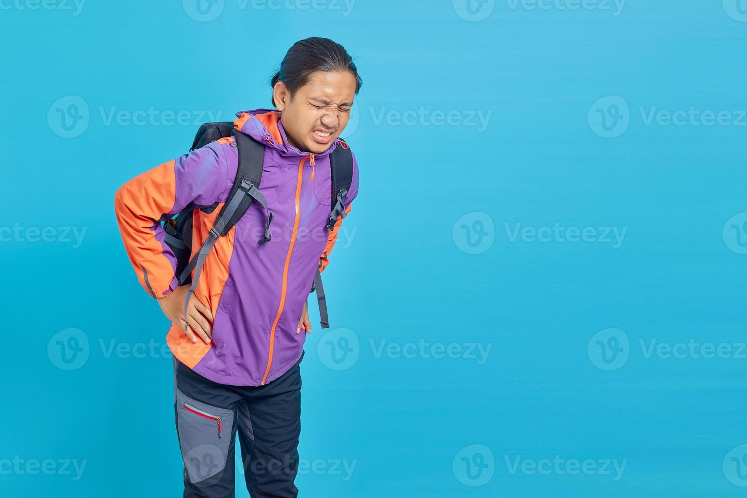 porträtt av stilig asiatisk man som lider av ryggsmärtor och röra ryggen med händerna på blå bakgrund foto