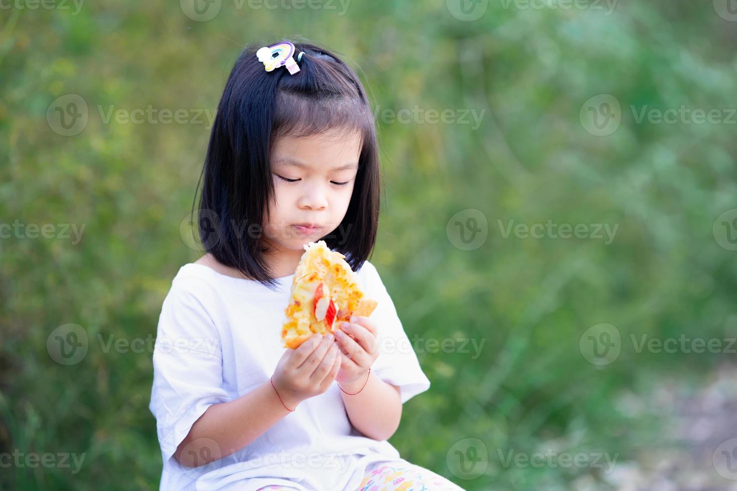 söt unge flicka äter pizza. bakom barnet var grön buske. foto
