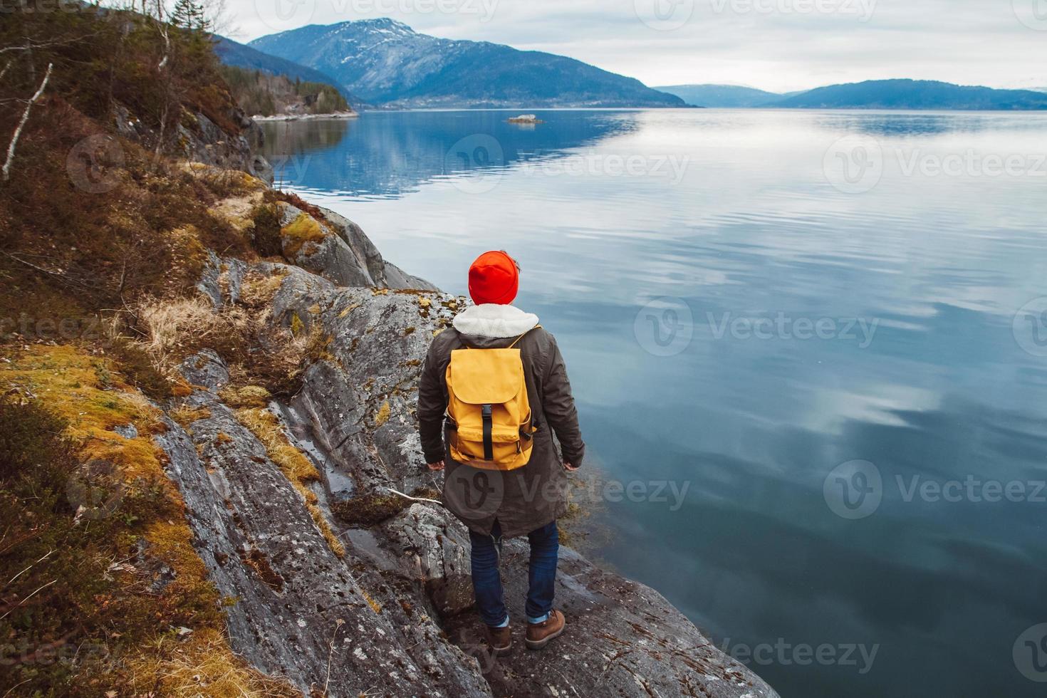 ung man med en gul ryggsäck som bär en röd hatt som står på en sten i bakgrunden av berg och sjö. utrymme för ditt textmeddelande eller reklaminnehåll. resa livsstilskoncept foto