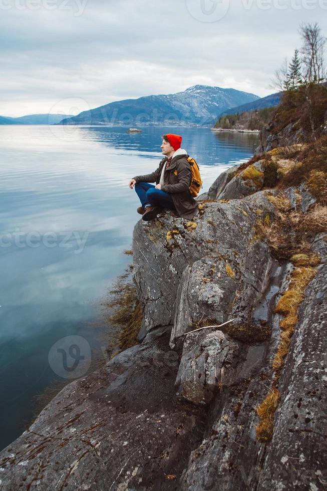 ung man med en gul ryggsäck som bär en röd hatt som sitter på stranden på bakgrund av berg och sjö. utrymme för ditt textmeddelande eller reklaminnehåll. resa livsstilskoncept. foto