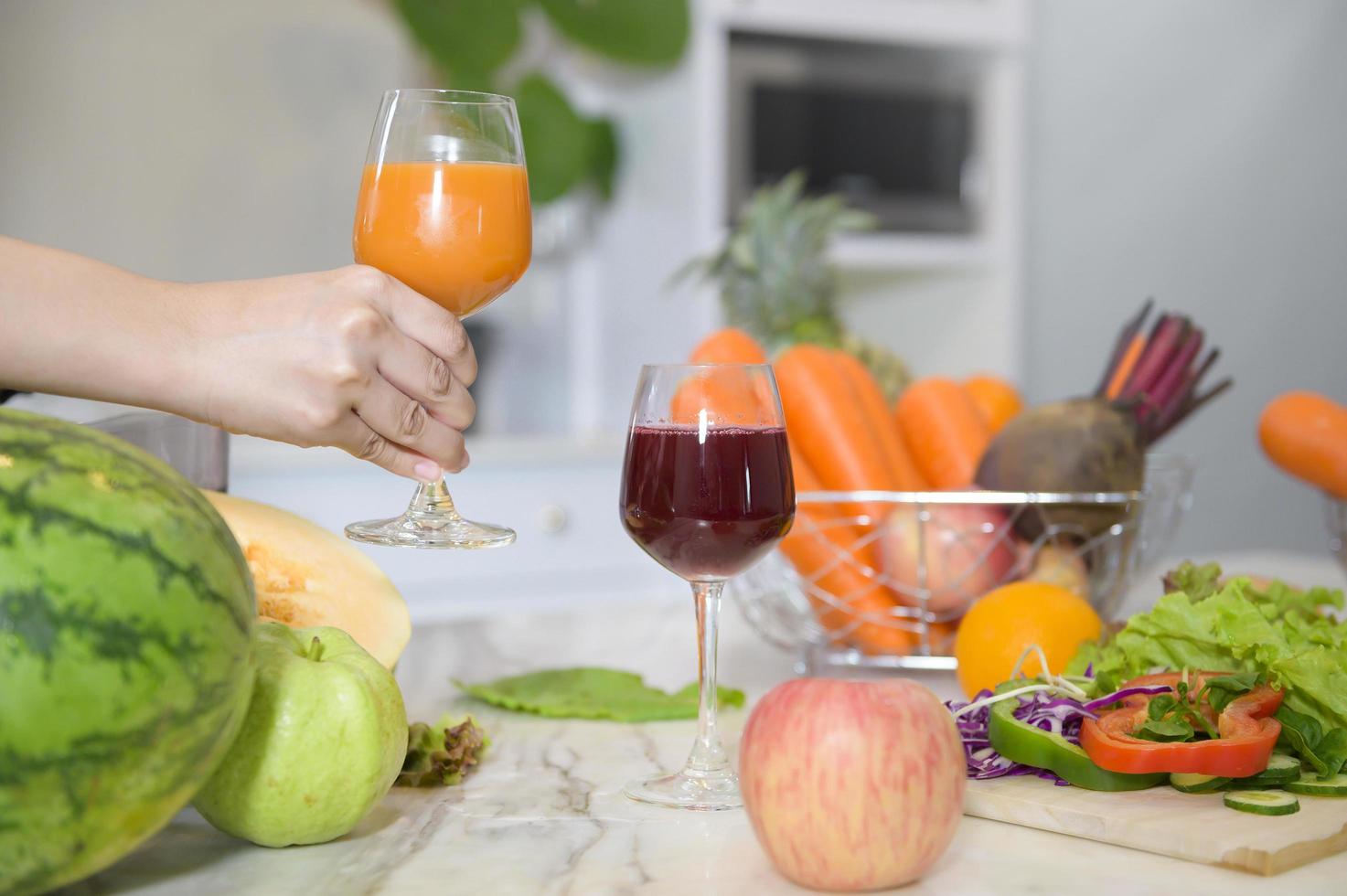 närbild av hand som håller ett glas hälsosam juice, medan grönsaker och juicepressar på bordet i köket, hälsokoncept foto