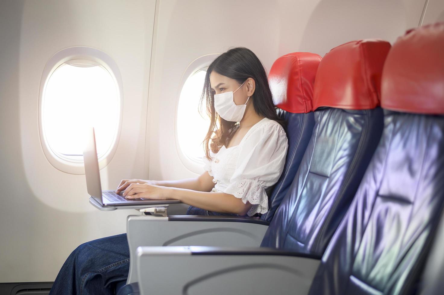 en ung kvinna som bär ansiktsmask använder bärbar dator ombord, ny normal resa efter covid-19 pandemikoncept foto