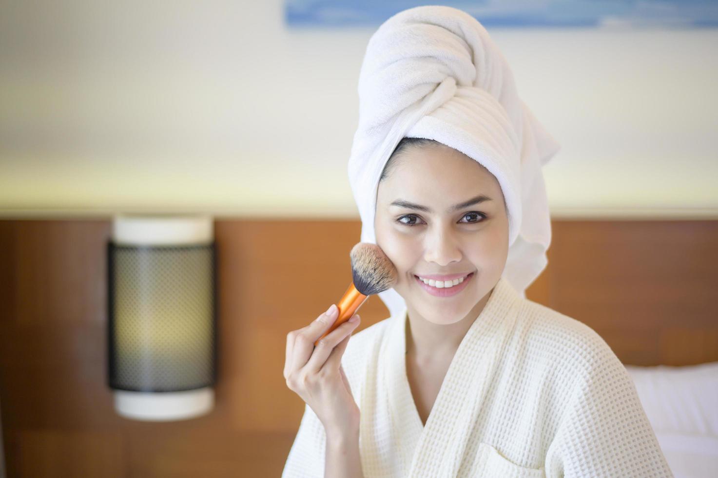 attraktiv glad kvinna i vit morgonrock applicerar naturlig make-up med kosmetisk puderborste, skönhetskoncept. foto
