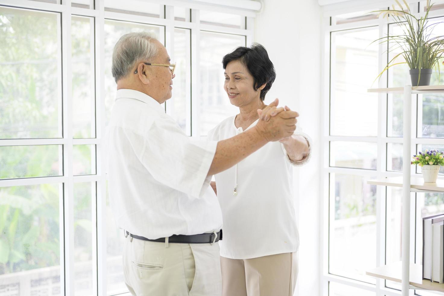 glada seniora asiatiska pensionerade par dansar och njuter av musiken i vardagsrummet hemma, hälsokoncept foto