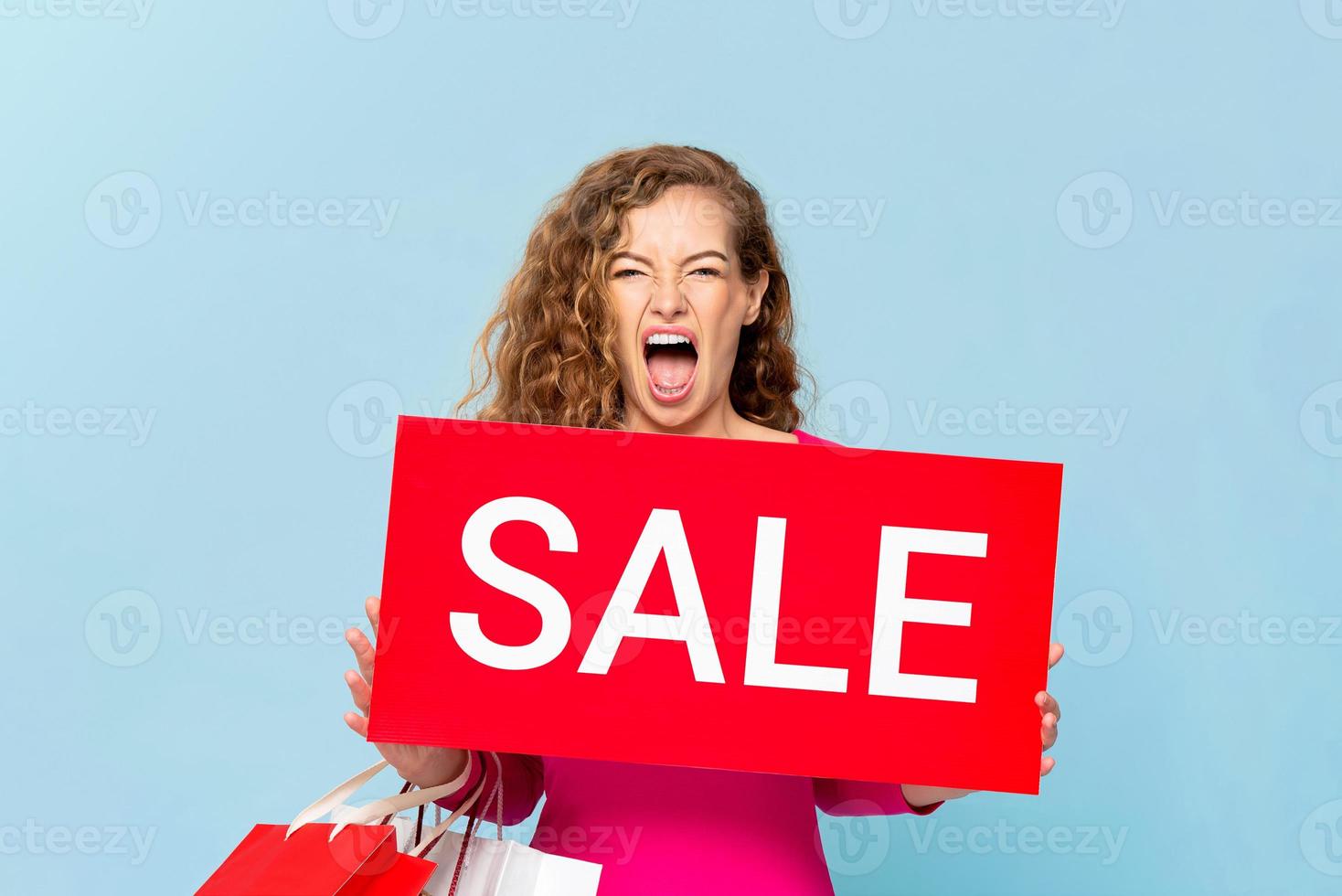 chockad kaukasisk kvinna som bär shoppingkassar visar röd försäljning skylt isolerad på ljusblå blå bakgrund foto