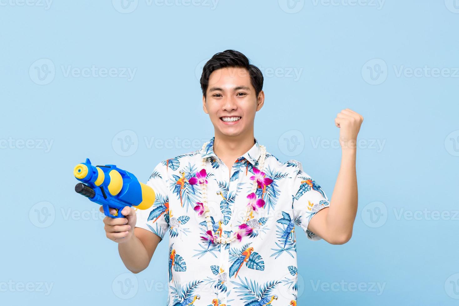 leende stilig asiatisk man som leker med vattenpistol och höjer näven isolerad på studioblå bakgrund för songkran-festivalen i Thailand och Sydostasien foto