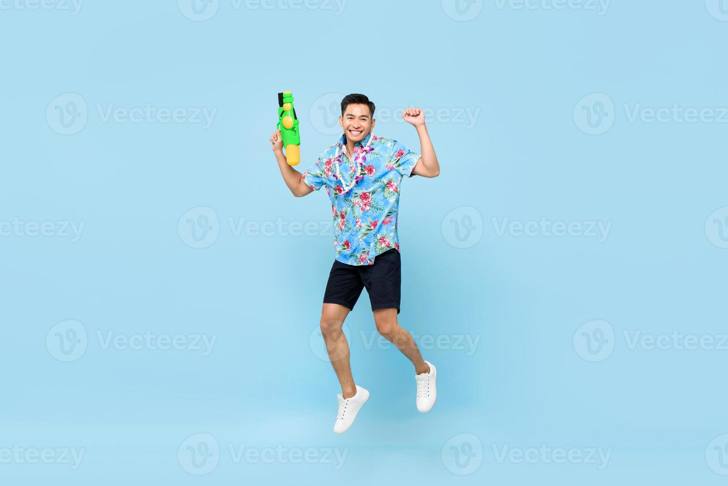 stilig leende ung asiatisk man som leker med vattenpistol och hoppar i studio blå bakgrund för songkran festival i Thailand och Sydostasien foto