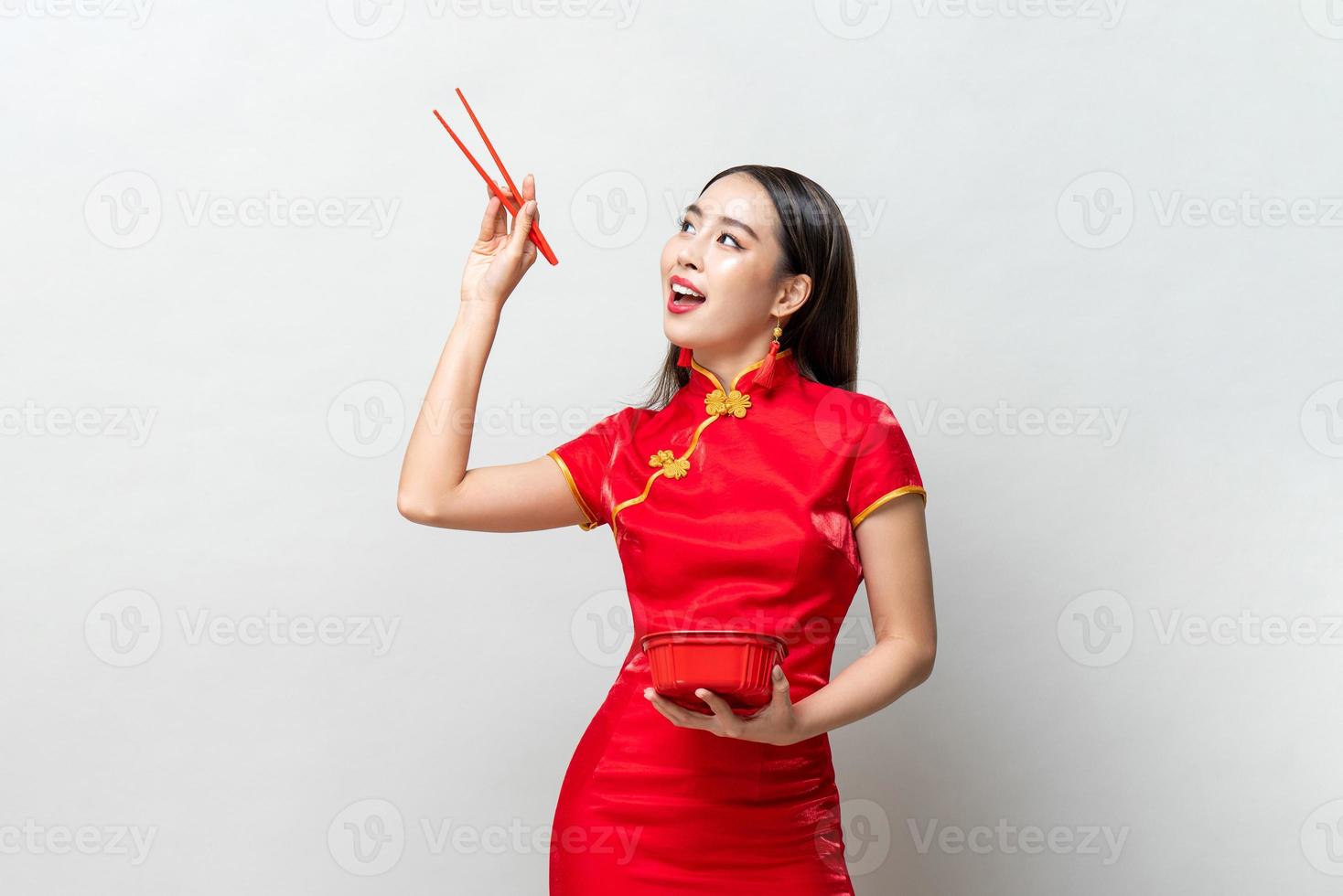 asiatisk kvinna i traditionell röd kinesisk qipaoklänning som håller plastmatlåda och ätpinnar tittar uppåt i studion isolerad ljusgrå bakgrund foto