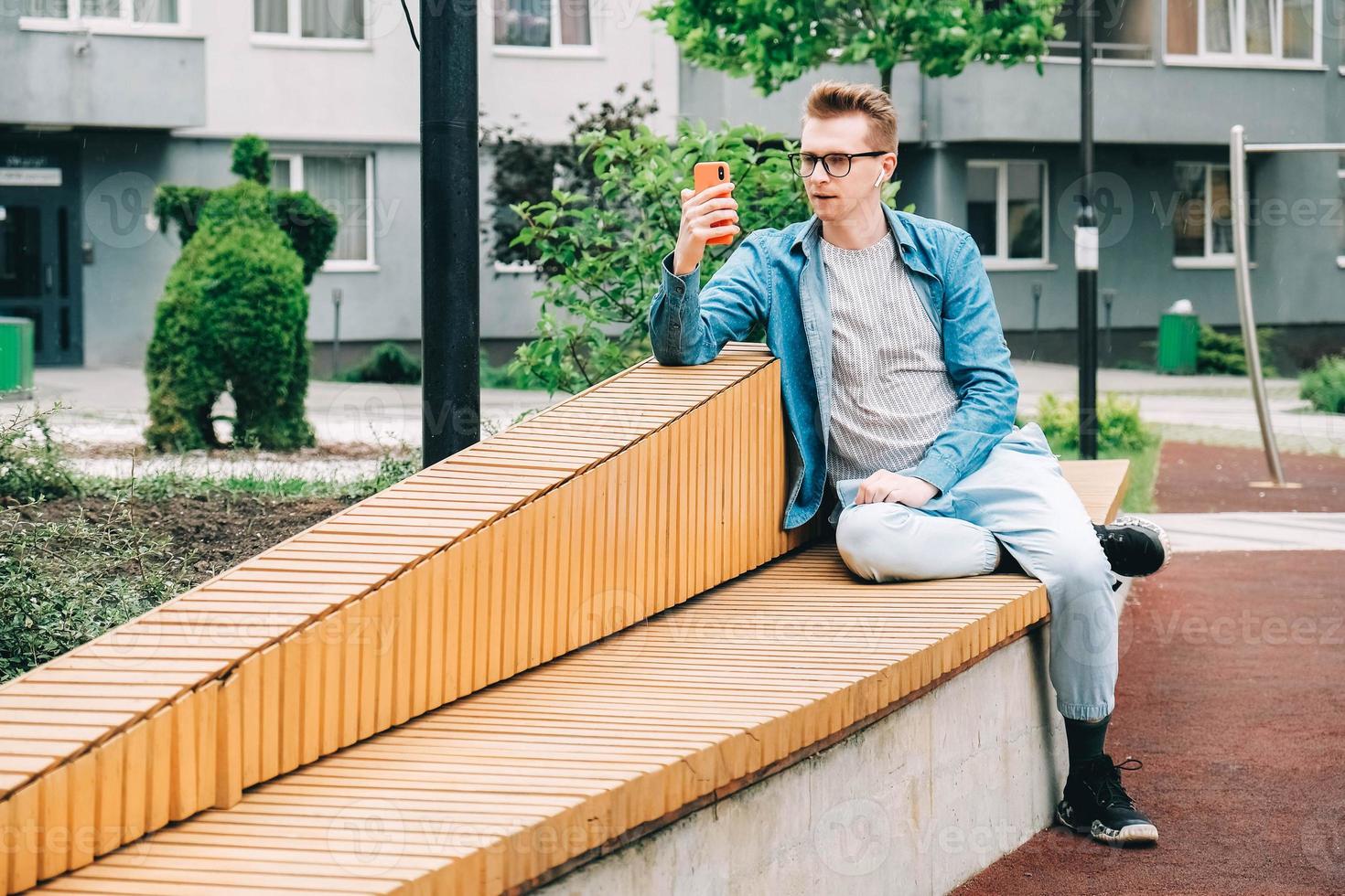 man i skjorta, jeans och glasögon som använder kommunicerar på en smartphone som sitter på en bänk på en bakgrund av hus foto