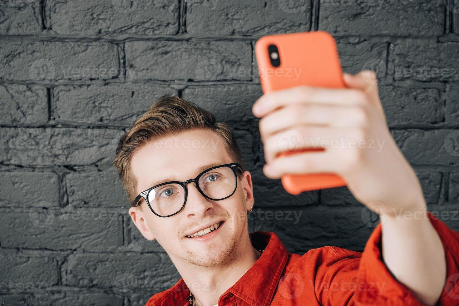 porträtt ung man i röd skjorta och glasögon tar selfiefoto på mobiltelefon på en bakgrund av svart tegelvägg. kopiera, tomt utrymme för text foto