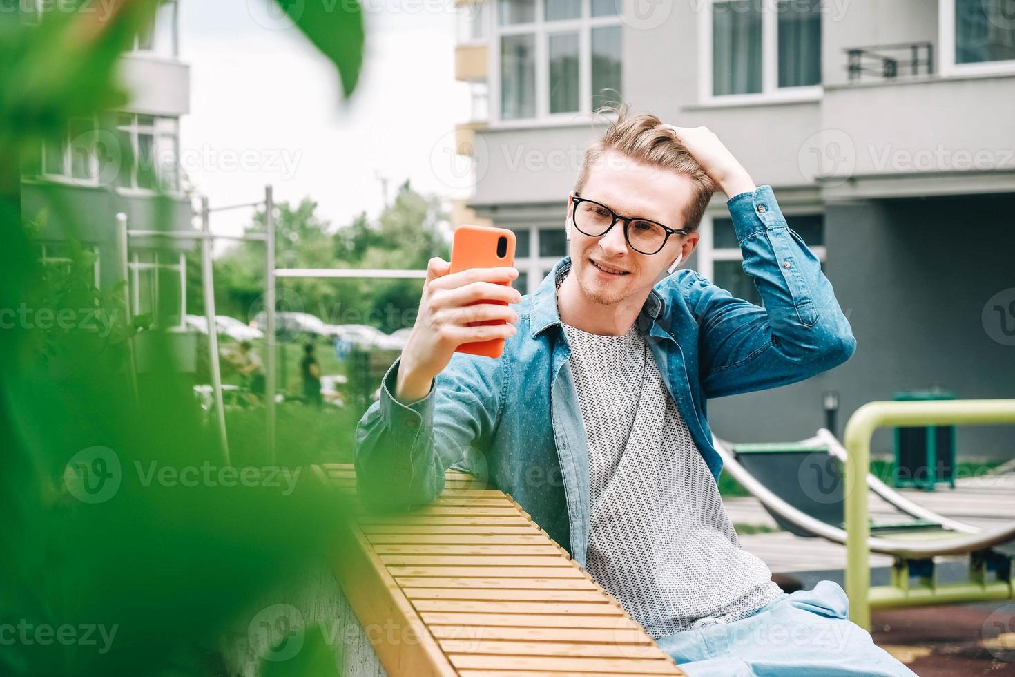 man i skjorta och glasögon använder kommunicerar på en smartphone som sitter på en bänk på en bakgrund av hus foto