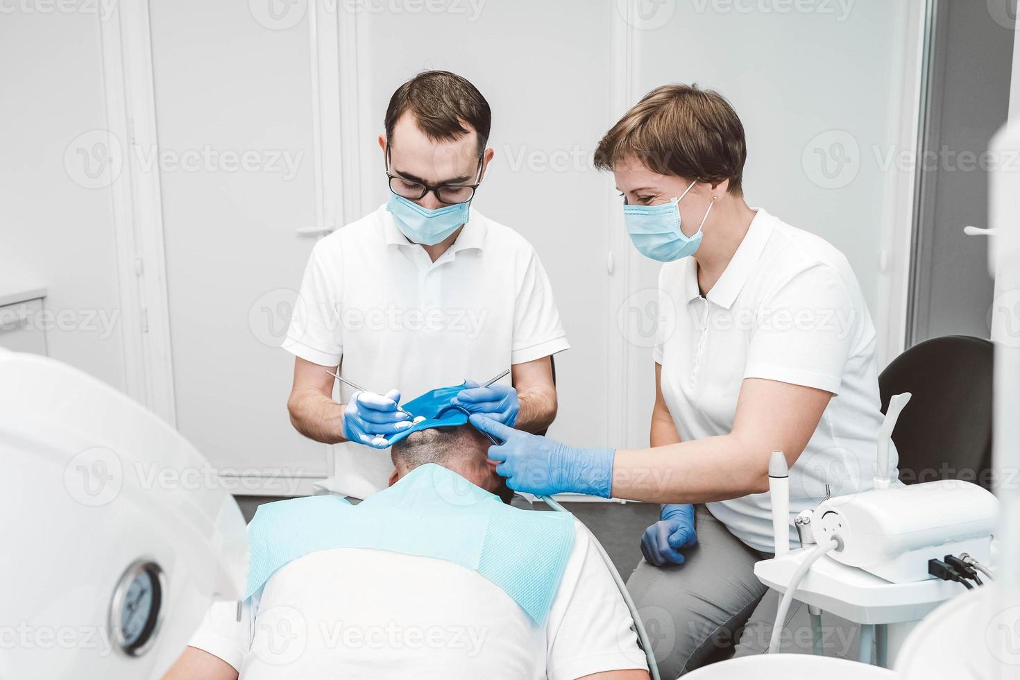 tandläkare och hans assistent behandlar en manlig patient på en tandvårdsklinik. tandläkare i medicinska masker som arbetar med klientens tänder foto