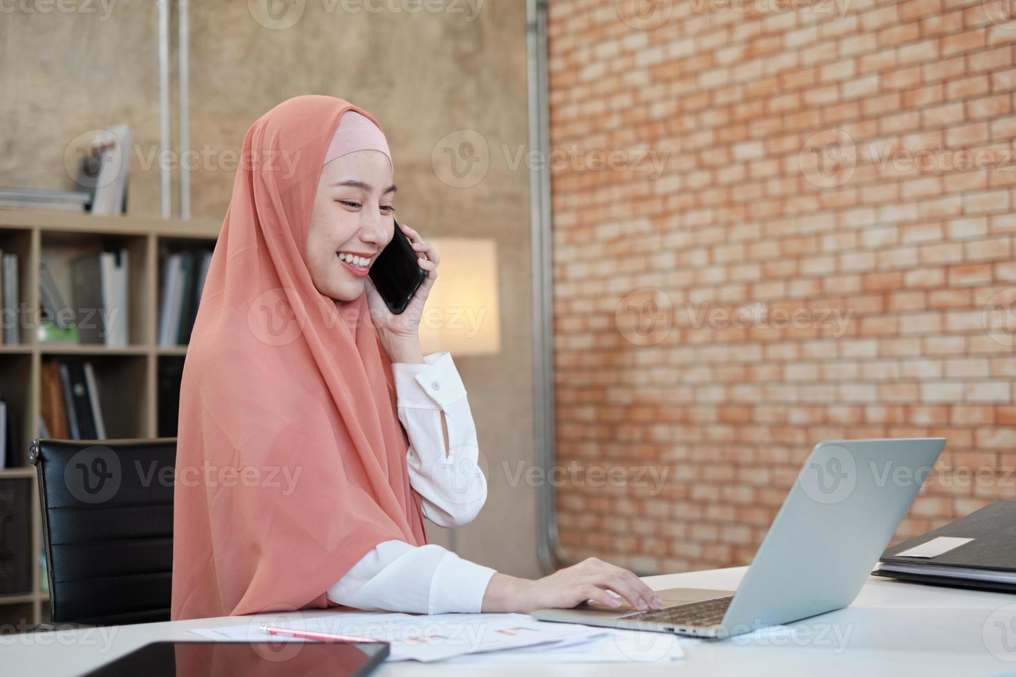 vacker affärskvinna som islamisk kvinnlig person pratar på sin mobiltelefon kommunikation och glada leende eftersom internet online beställning i laptop, modern livsstil, e-handel ockupation. foto