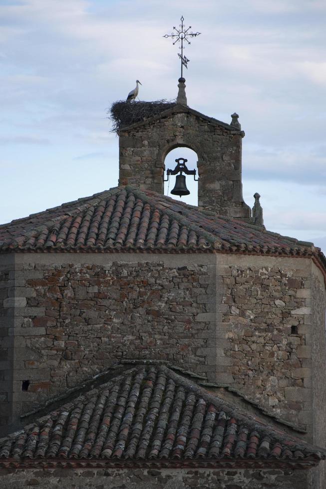 gammal lantlig kyrka i spanien gjord av sten. torn med en klocka, en metallisk vindmätare och en stork i sitt bo foto