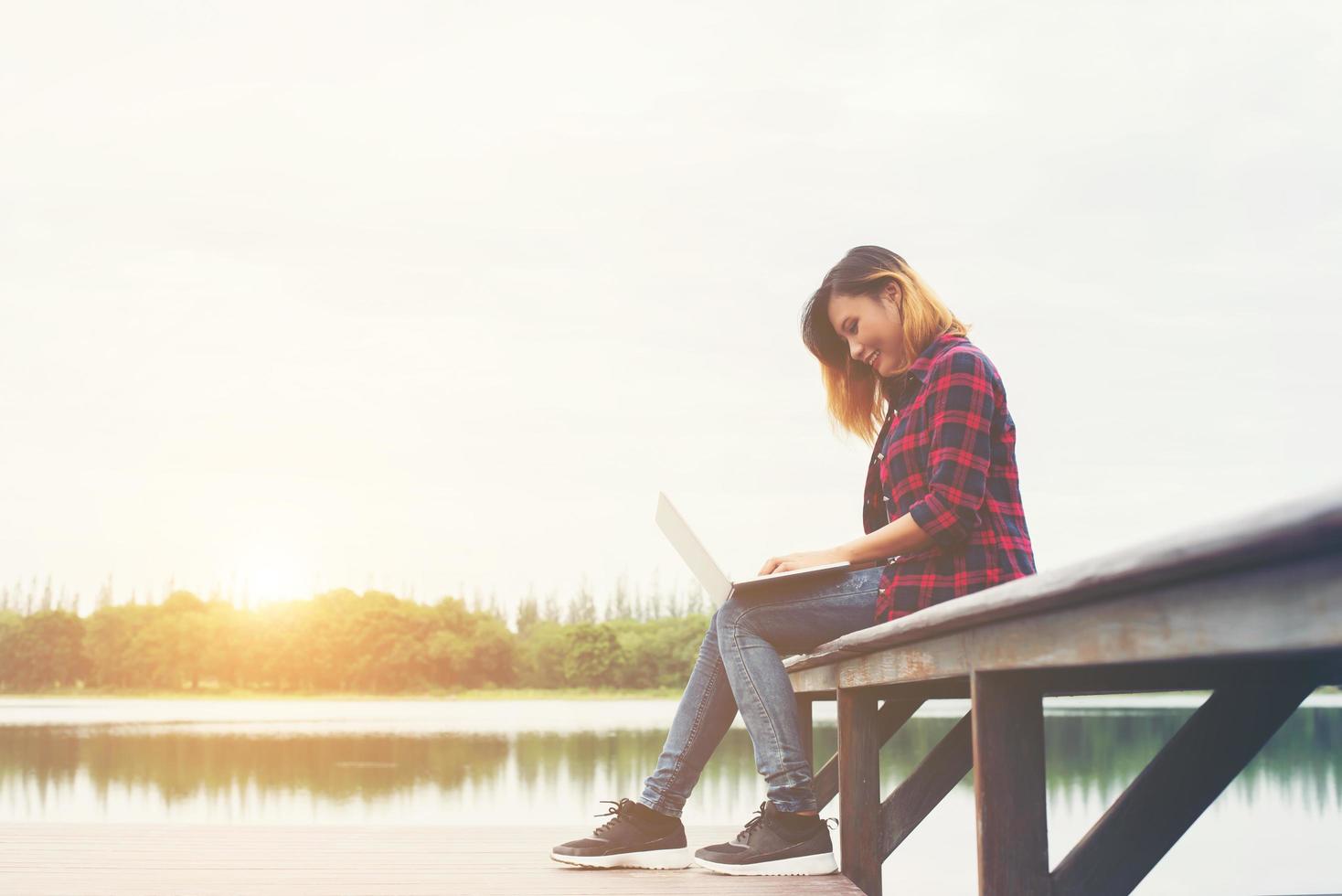 ung glad hipster kvinna som arbetar med sin bärbara dator sitter på piren, avkopplande njuta av naturen. foto