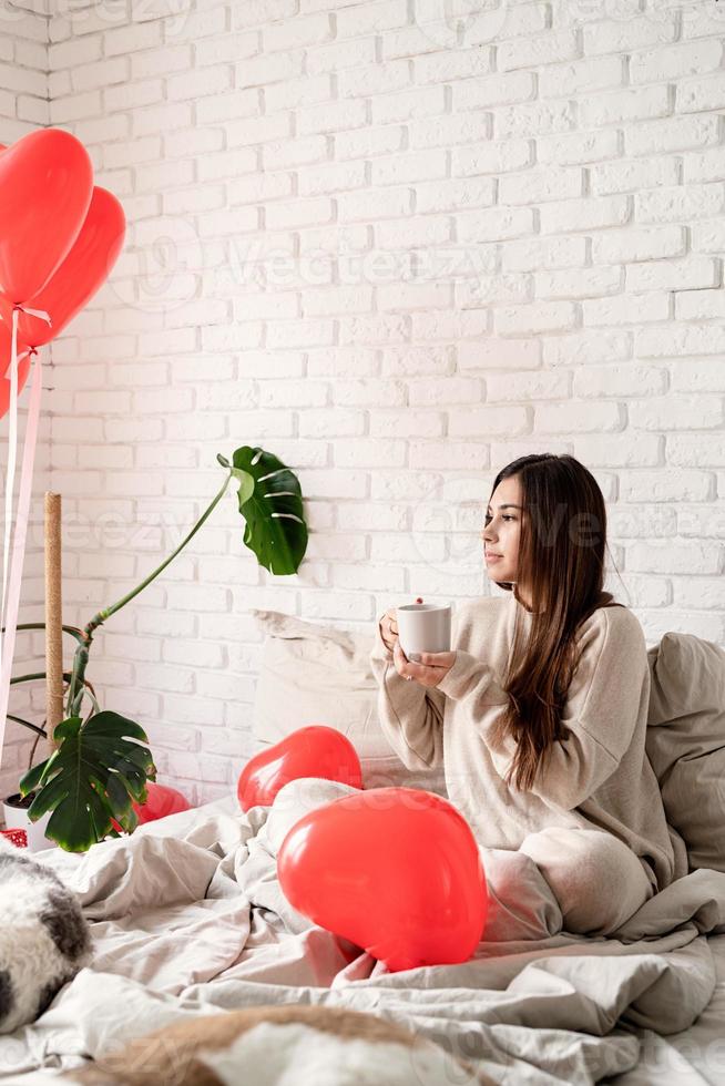 ung brunett kvinna sitter i sängen och firar alla hjärtans dag håller en kopp kaffe foto
