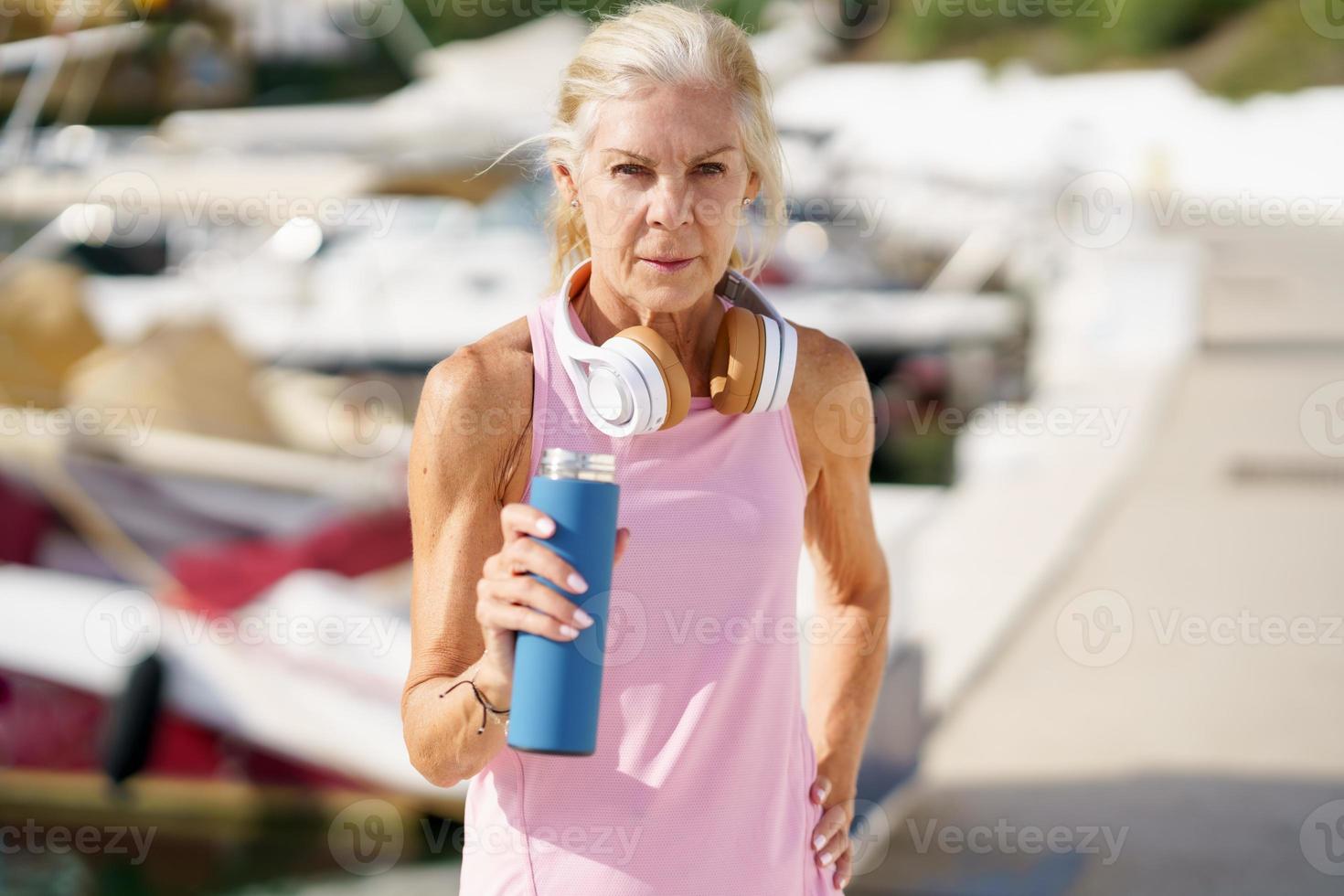 senior kvinna i fitness kläder dricksvatten från en metall fitness flaska utomhus. foto