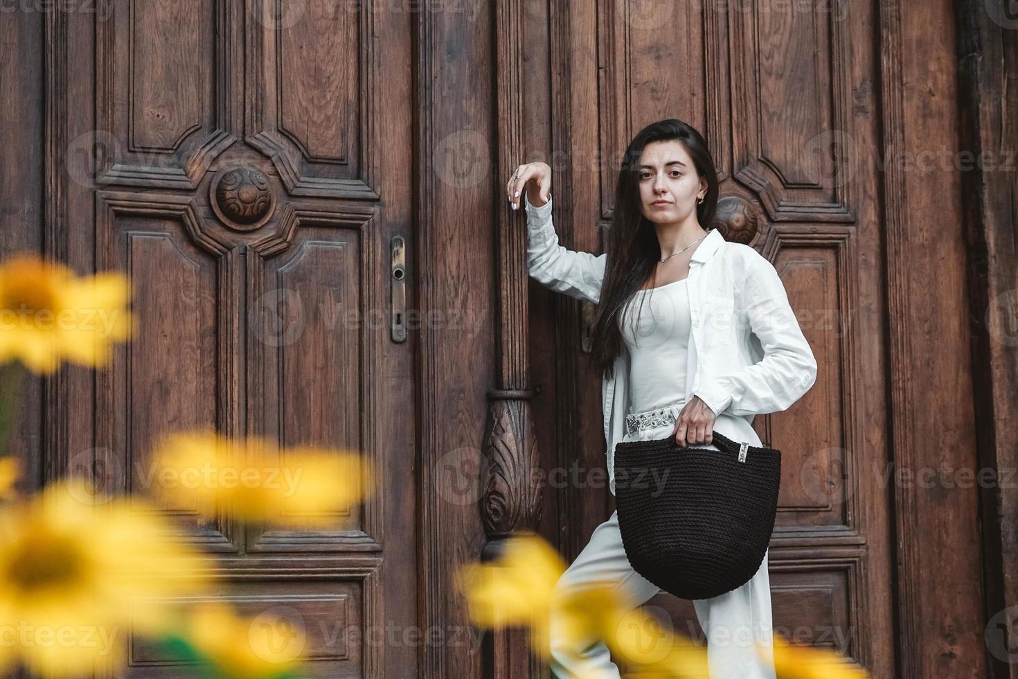 vacker ung brunett kvinna som bär i vita kläder, håller stickad väska på bakgrund av gamla trädörrar foto