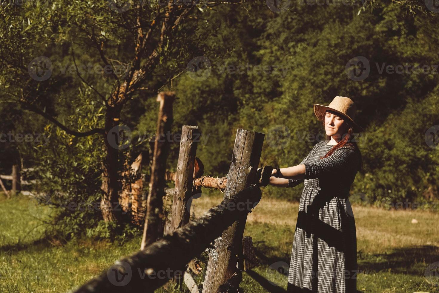 kvinna som bär solklänning och en stråhatt som står nära ett trästaket på en bakgrund av skog och träd vid solnedgången, med slutna ögon foto