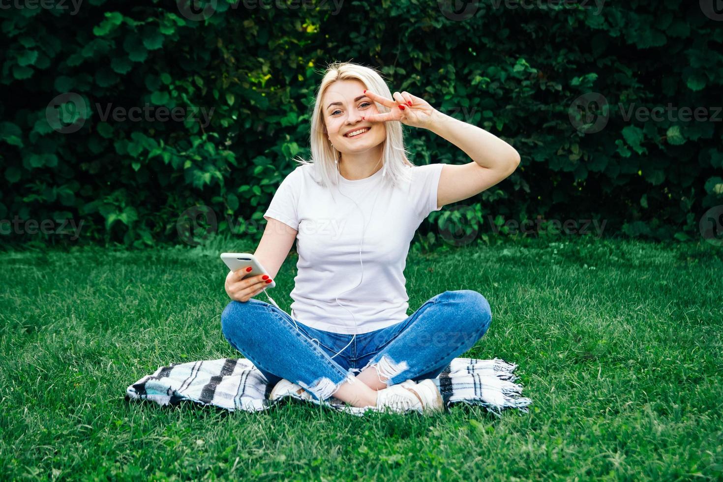 vacker blond kvinna i hörlurar och med en smartphone i händerna och lyssnar på musik som sitter på gräs utomhus i parken. plats för text eller reklam foto
