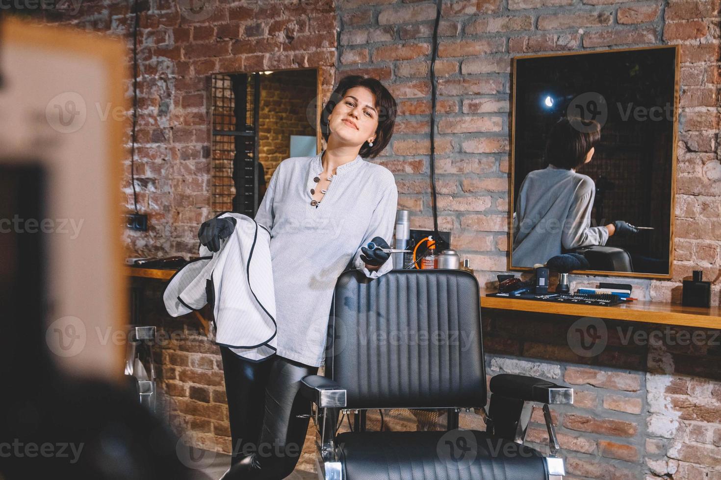 ung vacker frisör kvinna med sax i handen mot en tegelvägg bakgrund. barbershop reklam koncept. plats för text eller reklam foto