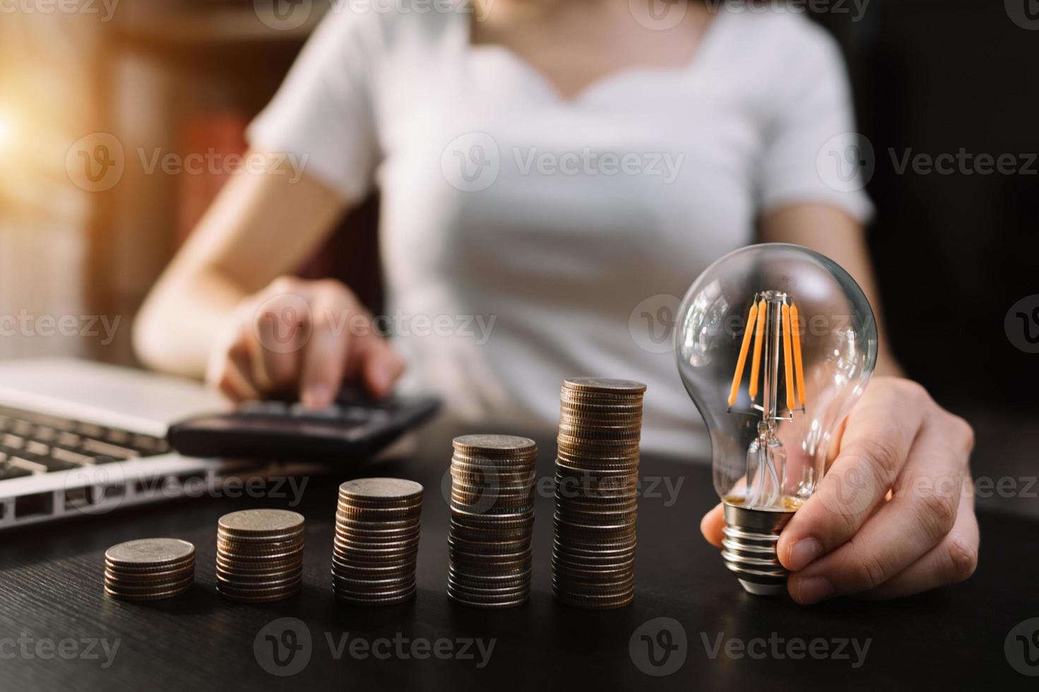 business hand som håller glödlampa med att använda bärbar dator och pengar stack på kontoret. idé att spara energi och redovisningsfinansieringskoncept i morgonljus foto