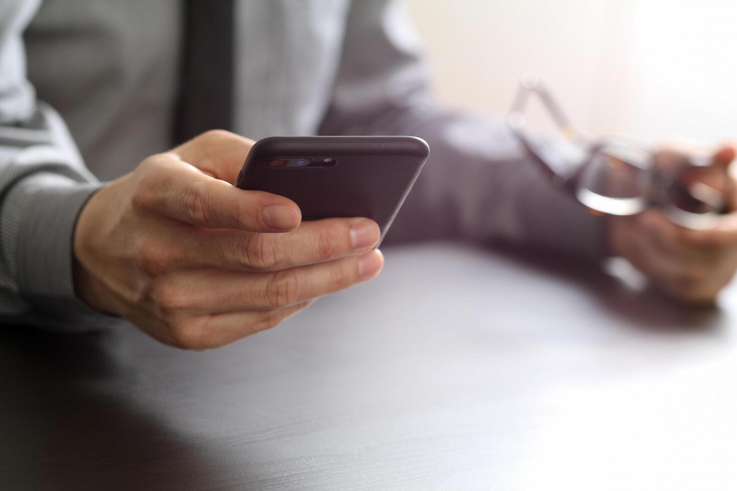 närbild av affärsman som arbetar med mobiltelefon och glasögon på trä skrivbord i moderna kontor foto