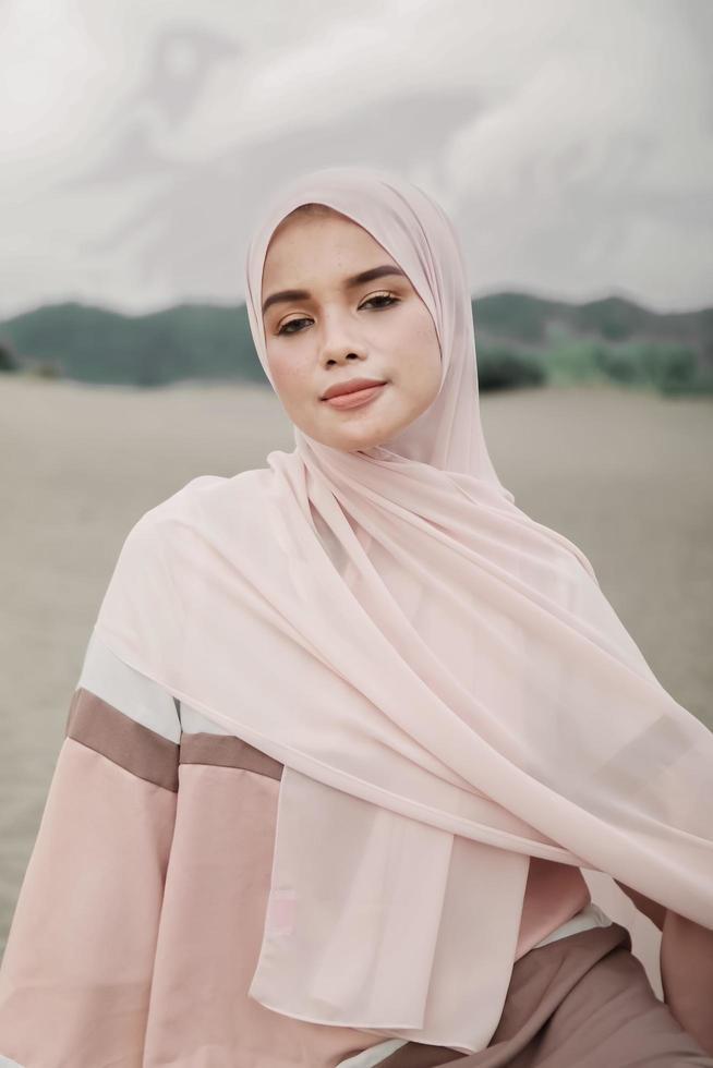 vacker islamisk kvinnlig modell som bär hijabmode, en modern bröllopsklänning för muslimsk kvinna som sitter i sanden och stranden. porträtt av en asiatisk tjejmodell med hijab som har kul på stranden med träd foto