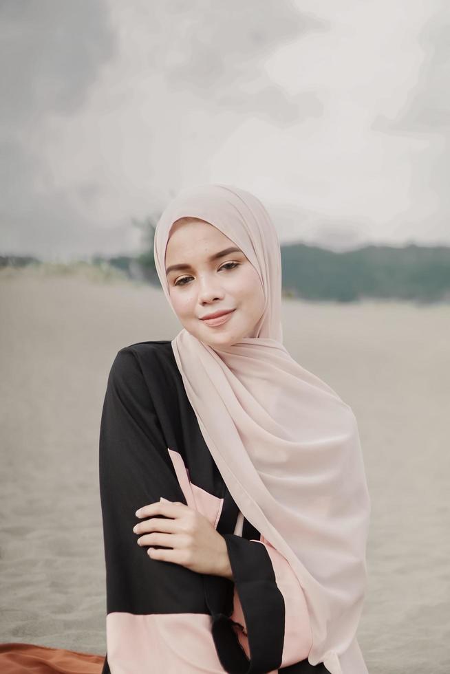 vacker islamisk kvinnlig modell som bär hijabmode, en modern bröllopsklänning för muslimsk kvinna som sitter i sanden och stranden. porträtt av en asiatisk tjejmodell med hijab som har kul på stranden med träd foto