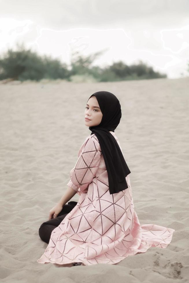 vacker islamisk kvinnlig modell som bär hijabmode, en modern bröllopsklänning för muslimsk kvinna som sitter i sanden och stranden. porträtt av en asiatisk tjejmodell med hijab som har kul på stranden med träd foto