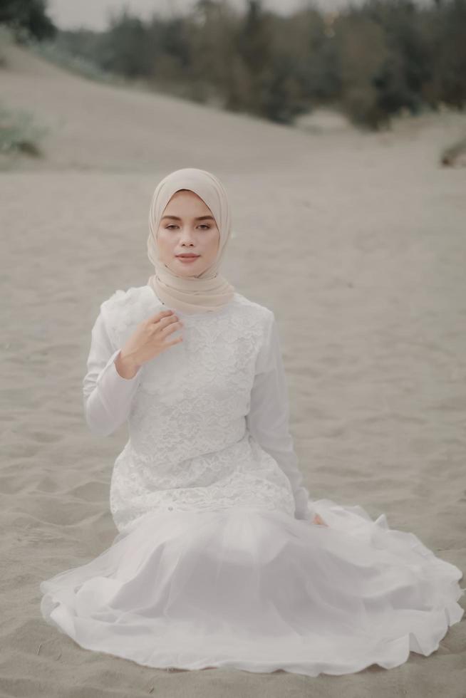 vacker islamisk kvinnlig modell som bär hijabmode, en modern bröllopsklänning för muslimsk kvinna som sitter i sanden och stranden. porträtt av en asiatisk tjejmodell med hijab som har kul på stranden med träd foto