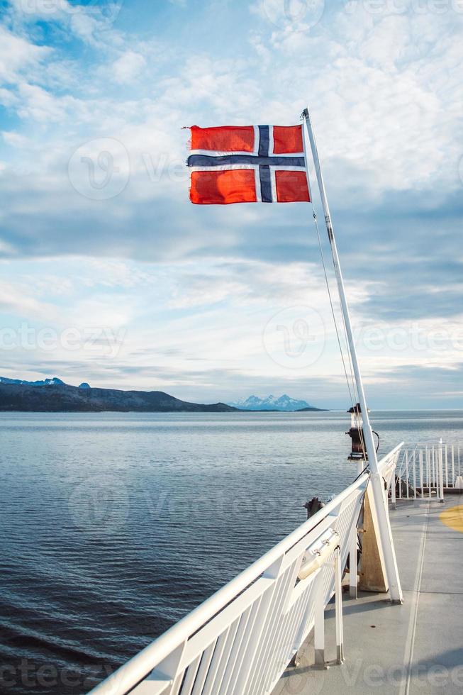 Norges flagga på havet och bergen bakgrund. norsk flagga hänger på fartygets reling och vajar ovanför vattnet. norska fjorden med en flagga foto