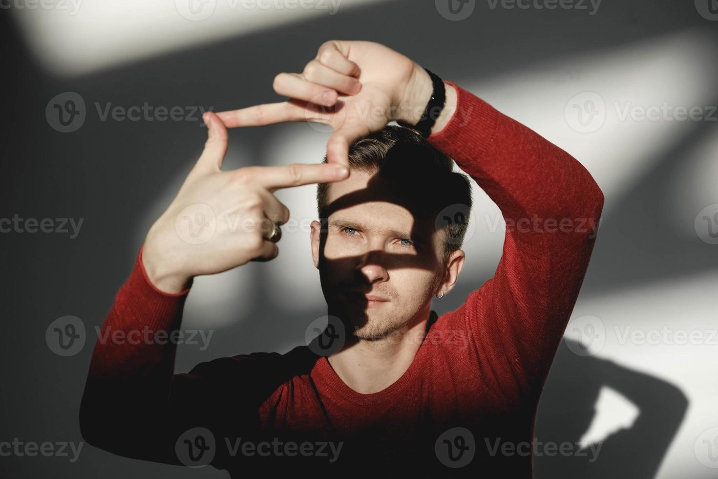 stilig man tittar genom en ram som bildas av hans händer i en röd tröja på en vit bakgrund med skuggor från ljuset. kopiera, tomt utrymme för text foto