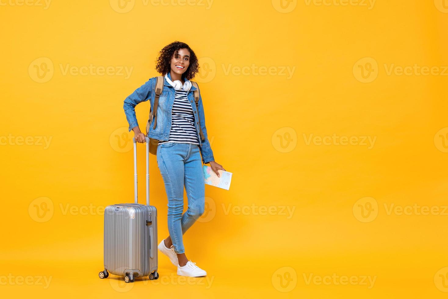 fullängds reseporträtt av leende ung afroamerikansk kvinna backpacker som står och håller bagage på gul studiobakgrund foto
