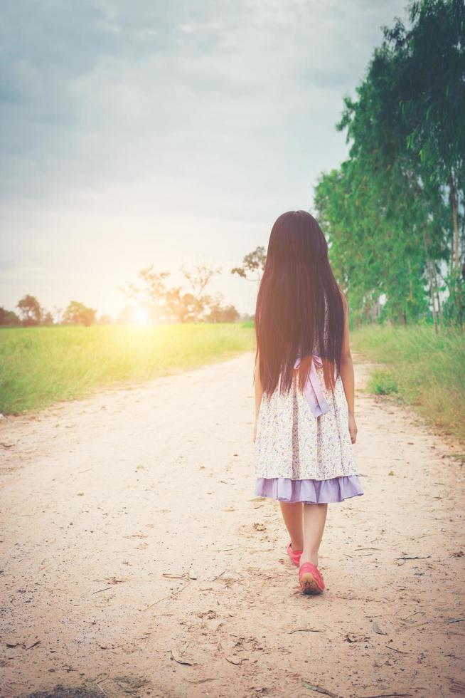 liten flicka med långt hår klädd i klänning går ifrån dig på landsväg. foto