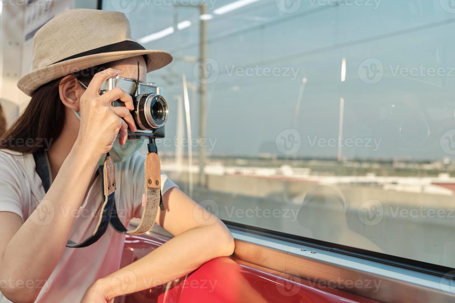 vacker asiatisk kvinnlig turist med ansiktsmask sitter i ett rött säte, reser med tåg, tar ögonblicksbilder, transporterar i förortsvy, njuter av passagerarlivsstil med järnväg, lycklig semester. foto
