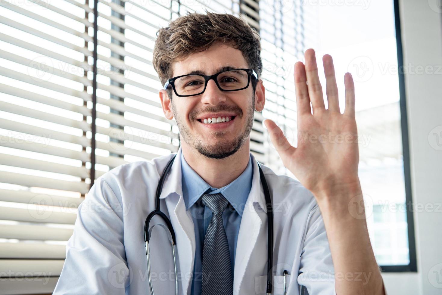 leende ung manlig läkare viftande hand till kameran hälsning patient online via distans videosamtal från kontoret foto