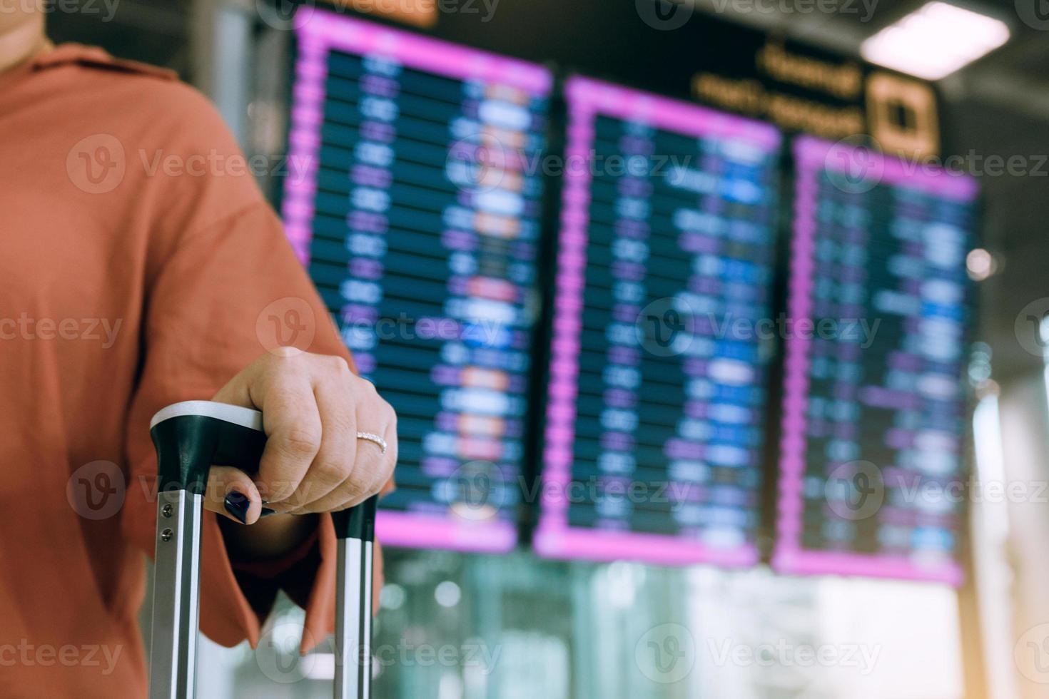 ung asiatisk kvinna som står på flygplatsen med resväska och kontrollerar avgång med flygschemat i bakgrunden. foto