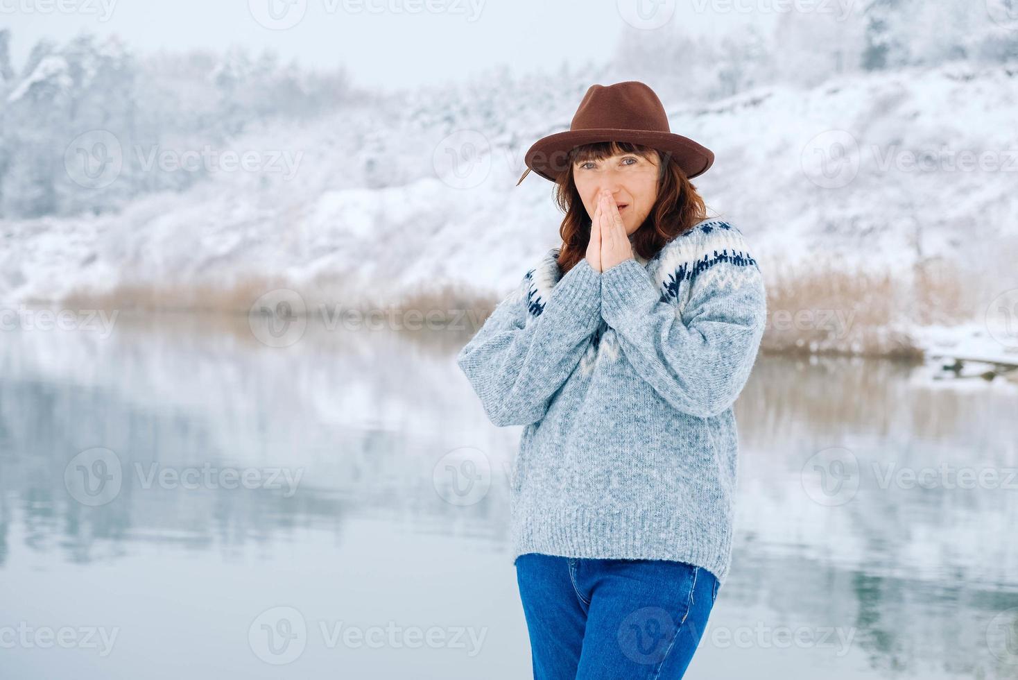 kvinna i brun hatt och tröja värmer handflatorna mot bakgrund av vinterskogen och sjön foto