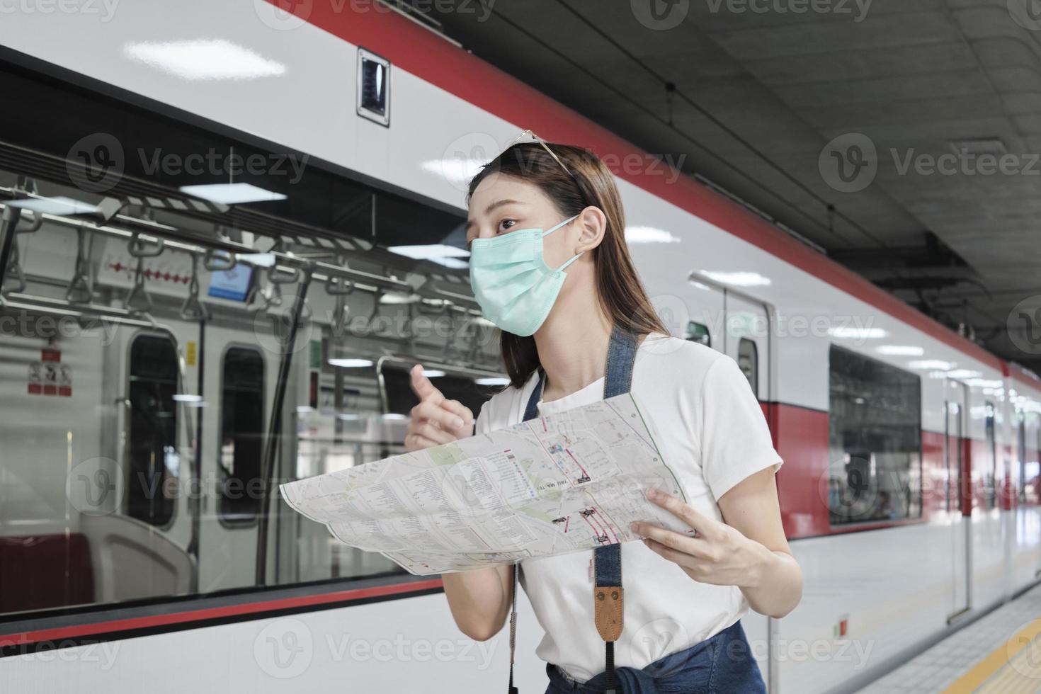 vacker asiatisk kvinnlig turist med ansiktsmask, glasögon och kamera, söker resenärens platskarta, letar efter destination på tågstationen i thailand, järnvägstransport, avslappnad livsstil. foto