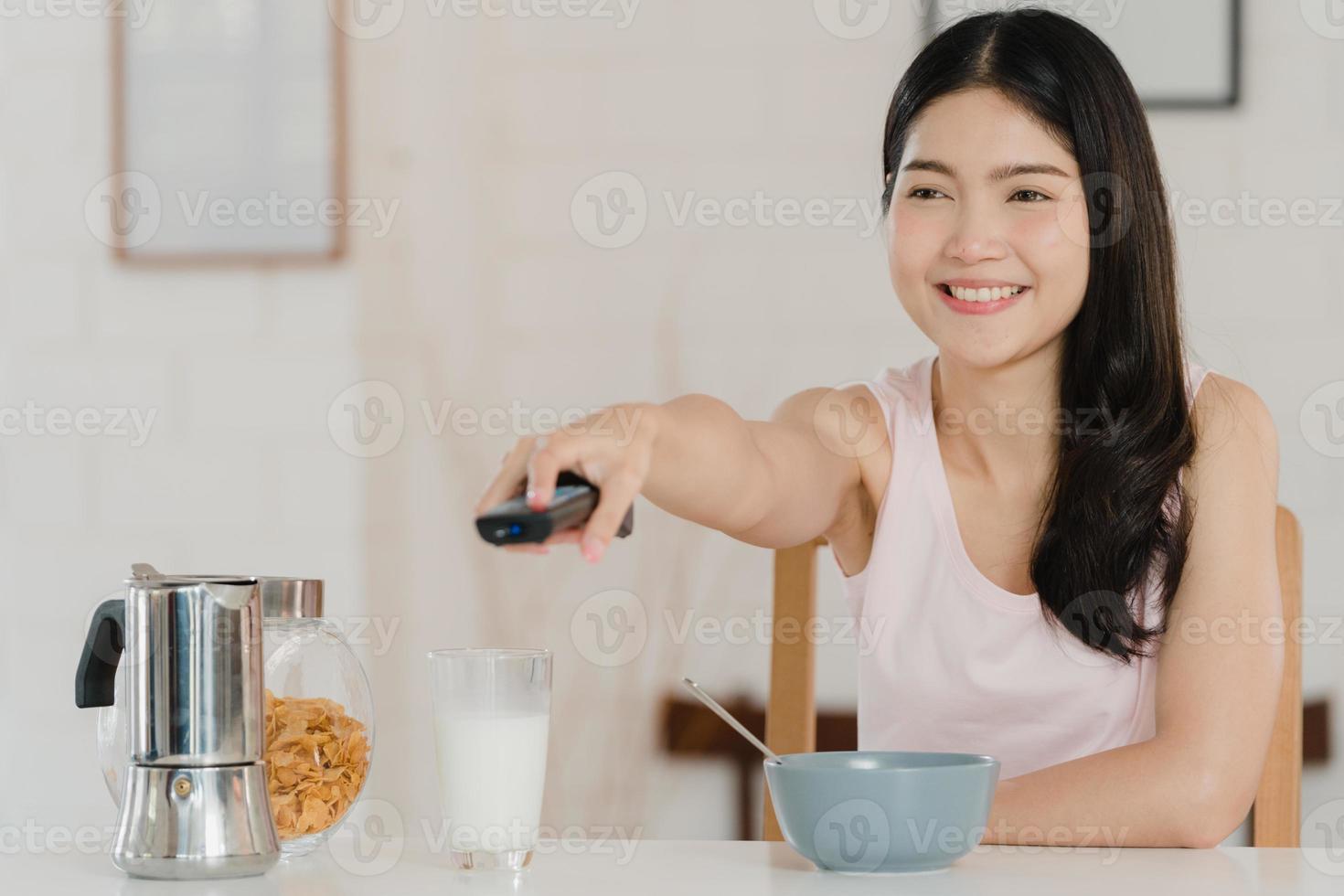 asiatisk japansk kvinna äter frukost hemma. unga asiatiska flickor känner sig glada när de tittar på tv medan de dricker juice, cornflakes spannmål och mjölk i skål på bordet i köket på morgonen koncept. foto