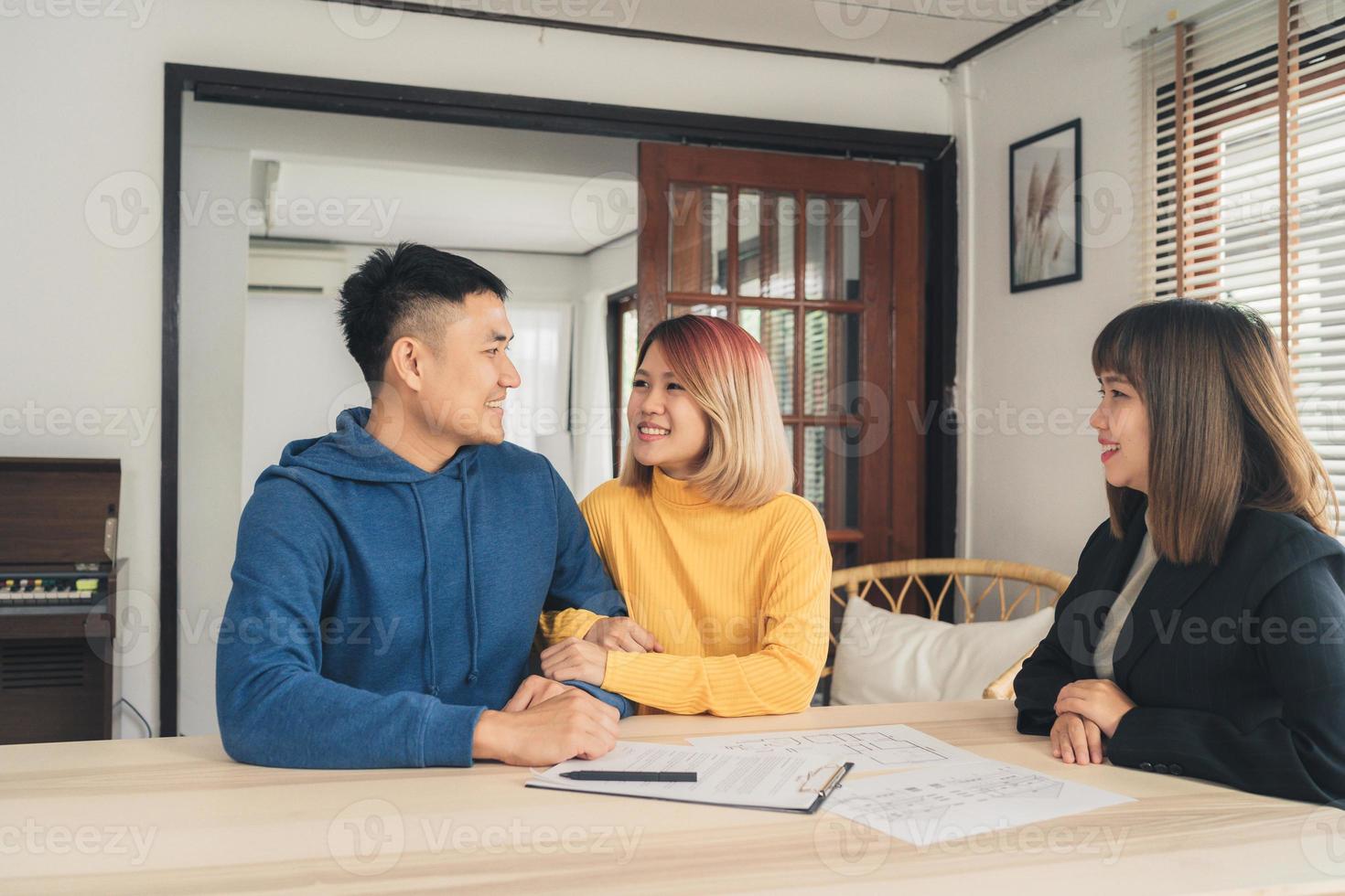 glada unga asiatiska par och fastighetsmäklare. glad ung man som undertecknar några dokument medan han sitter vid skrivbordet tillsammans med sin fru. köpa nytt hus fastigheter. undertecknar kontrakt i gott skick. foto