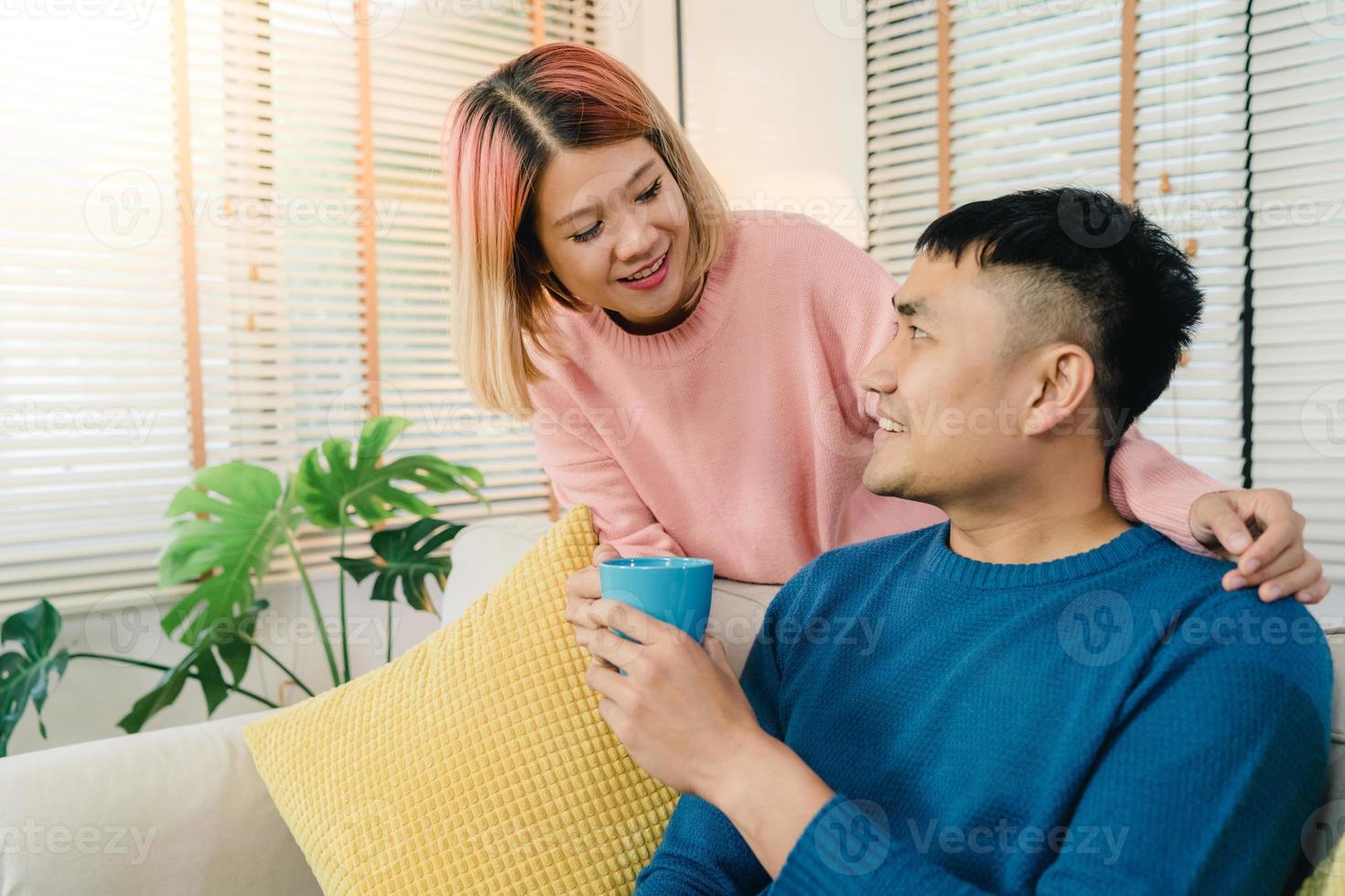 attraktiva asiatiska söta par njuta av kärleksögonblick dricka varm kopp kaffe eller te i händerna på soffan i vardagsrummet hemma. man och hans fru kopplar av hemma koncept. foto