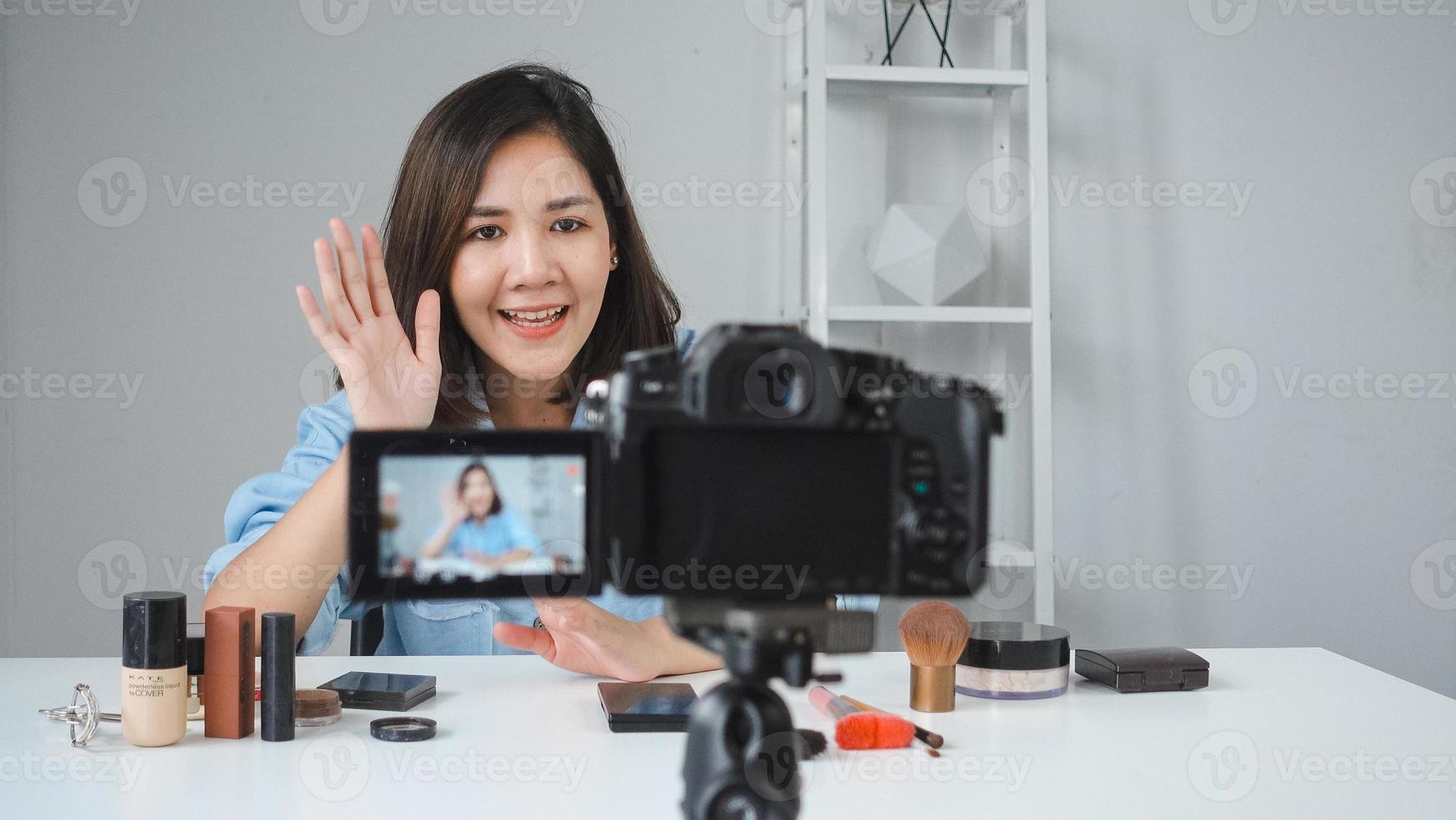 glad leende asiatisk kvinna eller skönhetsblogger med pensel och kamera som spelar in video och viftande hand hemma. skönhet videoblogg blogga människor koncept. foto