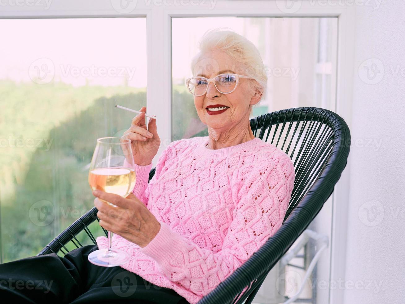 gammaldags senior snygg kvinna sitter på terrassen och röker cigarett med ett glas vitt vin. dålig vana, missbrukskoncept foto