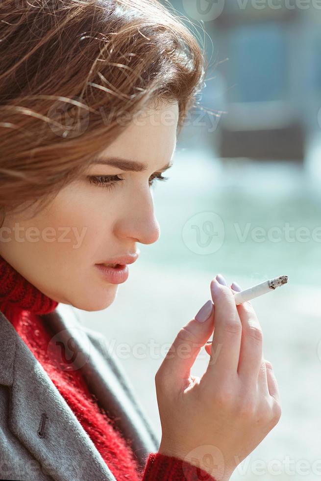 vacker snygg sorglig stressig ung kvinna i tröja och kappa röker cigarett nära floden foto