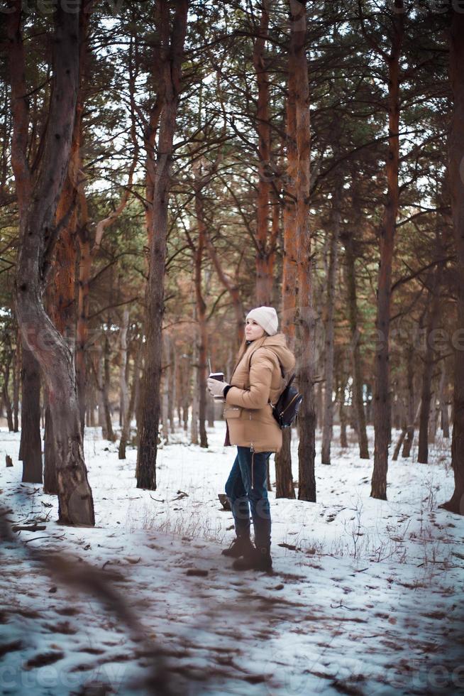 en attraktiv tjej med blont hår och en kopp varm dryck går i vinterskogen. foto